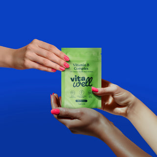 Two hands holding a green Vitawell vitamin package against a blue background
