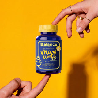 Hand holding a blue supplement bottle labeled 'Balance vitawell TEEN' against a yellow background
