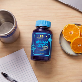 Vitawell Teen supplement bottle on a wooden table with a notebook, pen, and oranges.